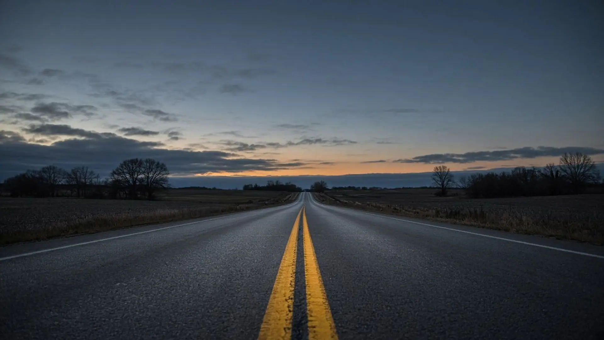 An empty two-lane country highway at dusk, its double yellow centerline running straight to the horizon between bare autumn fields.